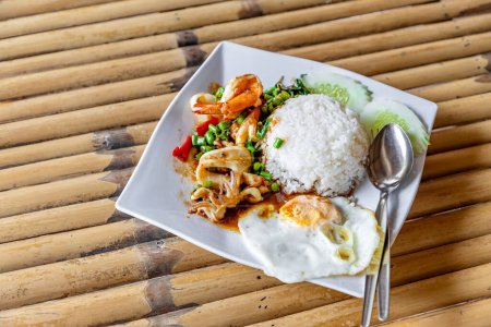 Foto de Comida tailandesa - Pad Kraprao Ta-le Khai Dao - Mariscos mezclados fritos con albahaca con un huevo frito en un plato blanco al lado, estilo tailandés. En una mesa hecha de bambú en un restaurante junto al mar. Durante el día. - Imagen libre de derechos