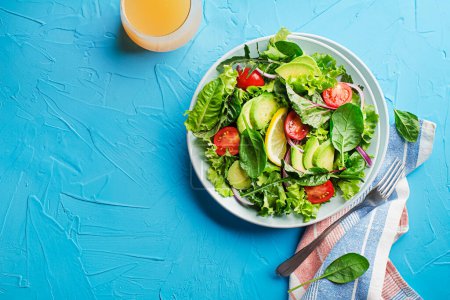Foto de Healthy green salad with avocado and fresh vegetables on blue table close up - Imagen libre de derechos