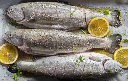 Foto de Fresh rainbow trouts with salt, lemon and herbs ingredients. Tasty fishes preparing for dish lunch. - Imagen libre de derechos
