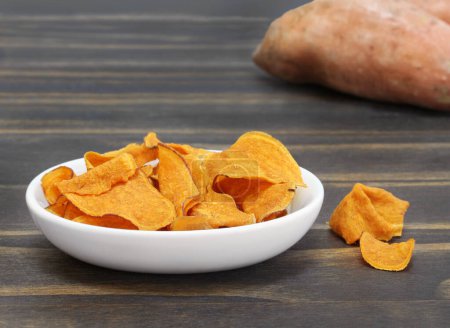 Photo for A bowl of sweet potato chips on a wooden table with a sweet potato in background.  Macro with copy space. - Royalty Free Image