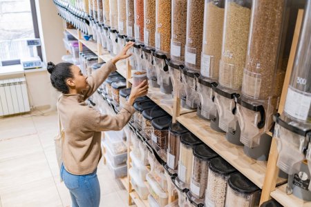 African American woman refilling reusable package with bulk products in local zero waste grocery store. Woman buying grains and cereals in sustainable eco grocery store. View from above