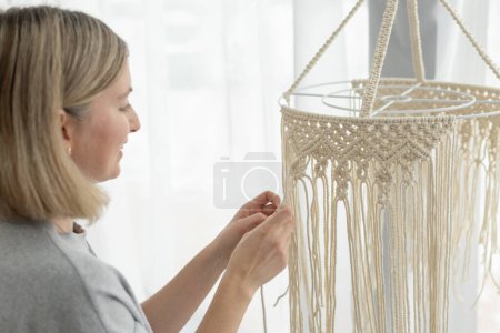 Eine Frau mittleren Alters strickt Lampenschirm für den Innenraum in Makrametechnik. Glückliche Frau strickt Boho-Kronleuchter