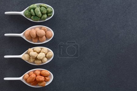 Photo for Variety of Japanese-style crunchy coated peanuts or cracker nuts with different flavoring (chives, original, chili) in small spoons, photographed overhead on slate with copy space on the side (Selective Focus) - Royalty Free Image