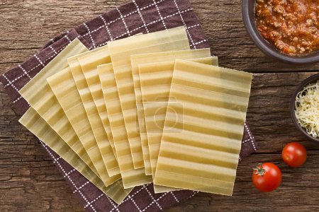 Photo for Dry uncooked ridged lasagna pasta sheets, photographed overhead on rustic wood with bolognese sauce, grated cheese and cherry tomato on the side - Royalty Free Image