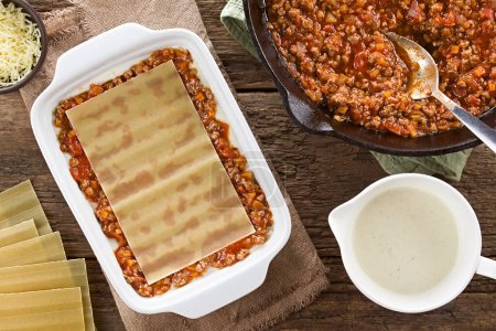 Photo for Preparing Lasagna in Rectangular Casserole Dish. Lasagna pasta sheet on top of the ragu or bolognese sauce and the white or bechamel sauce, ingredients on the side, photographed overhead on wooden table (Selective Focus, Focus on the lasagna pasta sh - Royalty Free Image
