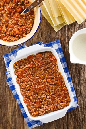 Photo for Preparing Lasagna in Rectangular Casserole Dish. Ragu or bolognese sauce on top of bechamel or white sauce, ingredients on the side, photographed overhead on wooden table (Selective Focus, Focus on the bolognese sauce in the casserole dish) - Royalty Free Image