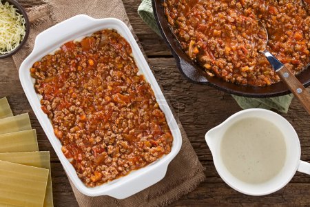Photo for Preparing Lasagna in Rectangular Casserole Dish. Ragu or bolognese sauce on top of bechamel or white  sauce, ingredients on the side, photographed overhead on wooden table (Selective Focus, Focus on the bolognese sauce in the casserole dish) - Royalty Free Image