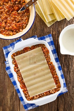 Photo for Preparing Lasagna in Rectangular Casserole Dish. Lasagna pasta sheet on top of ragu or bolognese sauce and bechamel or white sauce, ingredients on the side, photographed overhead on wooden table (Selective Focus, Focus on the lasagna pasta sheet in t - Royalty Free Image