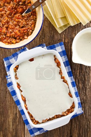 Photo for Preparing Lasagna in Rectangular Casserole Dish. Bechamel or white sauce on top of the ragu or bolognese sauce and the lasagna pasta sheet, ingredients on the side, photographed overhead on wooden table (Selective Focus, Focus on the white sauce in t - Royalty Free Image