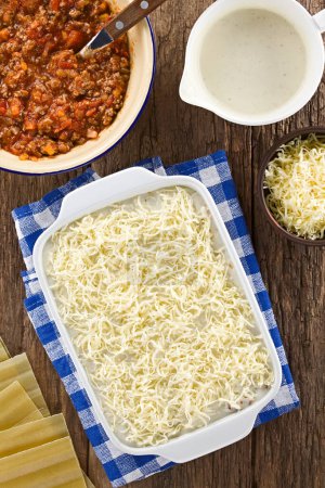 Photo for Grated cheese on top of the unbaked homemade lasagna, ingredients on the side, photographed overhead on wooden table (Selective Focus, Focus on the grated cheese in the casserole dish) - Royalty Free Image