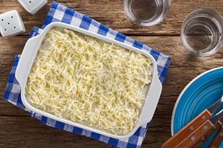 Photo for Unbaked homemade lasagna or casserole dish with grated cheese and white or bechamel sauce on top, glasses of water, plates with forks and salt and pepper shaker on the side, photographed overhead on wooden table (Selective Focus, Focus on the dish) - Royalty Free Image