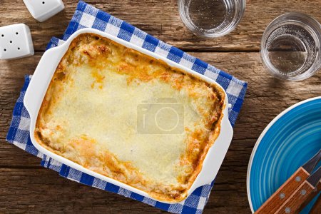 Photo for Freshly baked homemade lasagna or casserole dish with golden melted grated cheese on top, glasses of water, plates with forks, salt and pepper shaker on the side, photographed overhead on wooden table (Selective Focus, Focus on the dish) - Royalty Free Image