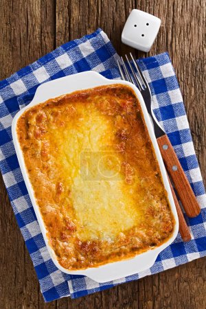 Photo for Freshly baked homemade lasagna or casserole dish with golden melted grated cheese on top, fork, knife and salt shaker on the side, photographed overhead on wooden table (Selective Focus, Focus on the dish) - Royalty Free Image