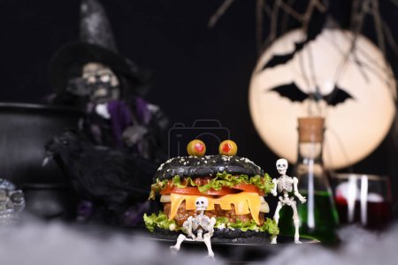 Foto de Hamburguesa monstruo. Pan negro, jugosa chuleta de ternera, lechuga, cebolla, tomate y queso en forma de dientes, en compañía de esqueletos. Definitivamente un pick-me-up y un aperitivo perfecto fiesta de Halloween - Imagen libre de derechos