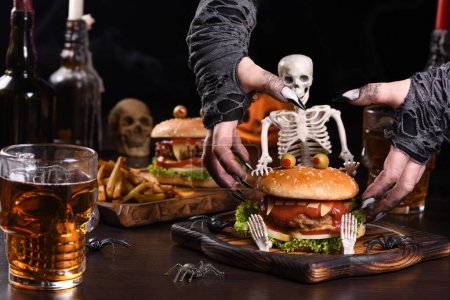 Foto de Las manos de brujas quieren agarrar la Hamburguesa Monstruo en el esqueleto sentado. Aperitivo de fiesta de Halloween perfecto - Imagen libre de derechos