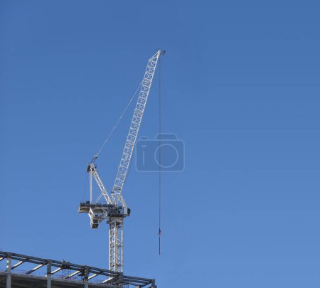 Tall hoisting tower crane on the top section of being constructed of modern high skyscraper building against clear blue cloudless sky