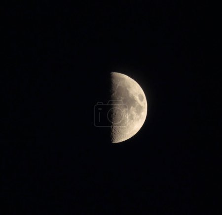 Half of Moon with craters on black night sky. Moon disk at moment of the first quarter phase, zoom view on a telescope.