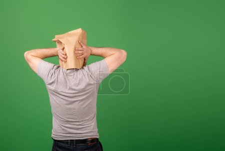 Ein unkenntlich gemachter Mann mit einer Papiertüte auf dem Kopf packte seinen Kopf vor einem grünen Hintergrund. Platz für Ihren Text. Psychische Gesundheit, Greenwashing, Desinformationskonzept