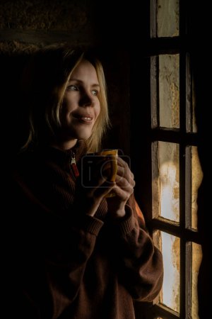 Téléchargez les photos : A woman smiles and drinks coffee by the window in the dark. A woman in her forties. - en image libre de droit