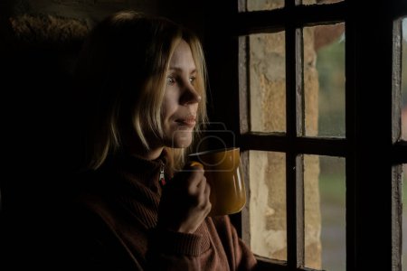 Téléchargez les photos : A woman drinks coffee in the dark by the window. A woman in her forties. Thoughtfulness, contemplation. - en image libre de droit