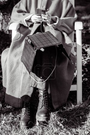Photo for An unrecognizable woman with a mobile phone in her hands and a portable solar battery sits outdoors in the garden. Ecological alternative energy concept. black and white photo. - Royalty Free Image