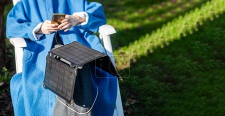 Photo for An unrecognizable woman with a mobile phone in her hands and a portable solar battery sits outdoors in the garden. Ecological alternative energy concept. - Royalty Free Image