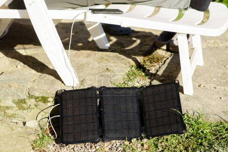 Photo for The solar battery lies on the ground near the plastic chair on which the mobile phone is charged. Alternative energy source concept. - Royalty Free Image