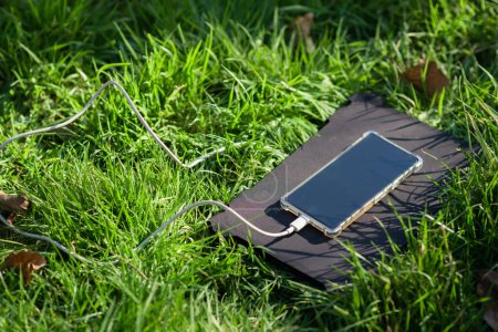 Photo for Portable solar battery and mobile phone on green grass. Charging electronic gadgets on a hike, during outdoor recreation. Ecological energy concept. - Royalty Free Image