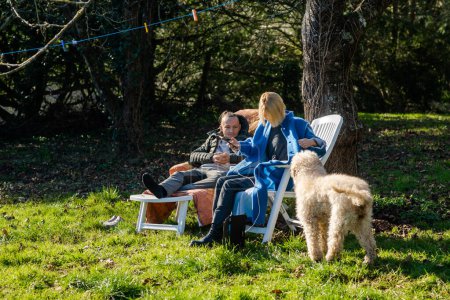 Photo for A man and a woman and a dog sit in the spring on sun loungers in the garden on green grass. - Royalty Free Image