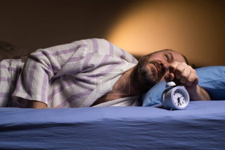 Foto de Reloj despertador para un hombre que se acuesta en la cama y sonríe. Facilidad de despertar, buen sueño, el modo adecuado para. - Imagen libre de derechos