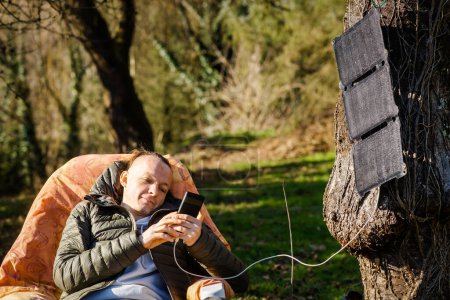 Photo for A portable solar battery is hanging from a tree. A man resting in nature with a mobile phone in his hands. Charging phone from solar eco energy. - Royalty Free Image