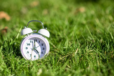 Foto de Reloj despertador de color blanco sobre hierba verde. Lugar para el texto. Tiempo, ritmo circadiano, concepto de madrugada. - Imagen libre de derechos