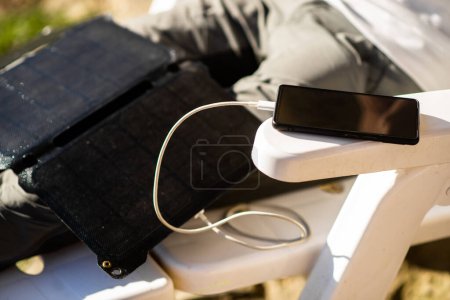 Photo for Portable solar battery. Lying on the legs of a resting unrecognizable man. The mobile phone is charged by a solar battery. Ecological energy. - Royalty Free Image