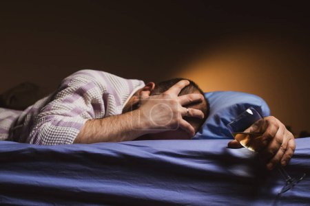 Foto de A man with a headache holds a glass of wine in bed. Alcoholism, loneliness, hangover concept. A dark room. - Imagen libre de derechos
