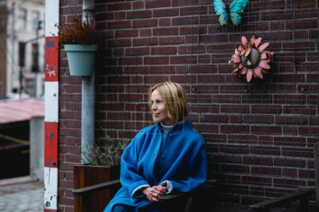 Téléchargez les photos : Une jolie femme adulte en manteau bleu regarde sur le côté. Derrière elle se trouve un mur de briques. Du beau temps. Voyage et aventure. - en image libre de droit