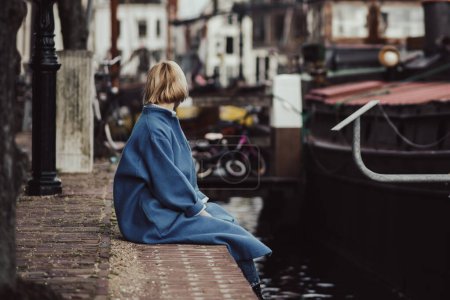 Téléchargez les photos : Une femme méconnaissable dans un élégant manteau bleu est assise sur la jetée. Zone résidentielle aux Pays-Bas. tonique. - en image libre de droit