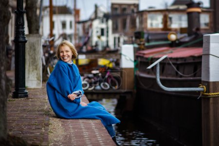 Téléchargez les photos : Une femme adulte heureuse et satisfaite, vêtue d'un élégant manteau bleu, est assise sur un quai. Quartier résidentiel aux Pays-Bas. - en image libre de droit