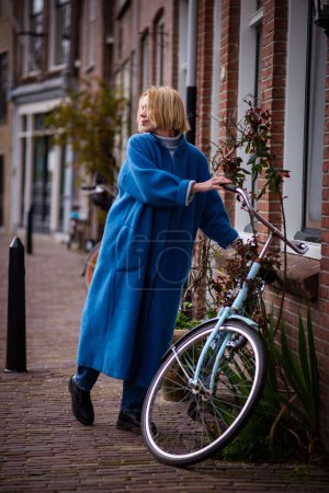 Téléchargez les photos : Une femme en long manteau bleu aux Pays-Bas. Touriste en voyage à Rotterdam. Une femme adulte prend un vélo. - en image libre de droit