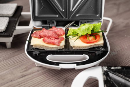 Foto de Una sandwichera con dos sándwiches diferentes con tomates, jamón, cebolla y queso. Preparación de una comida ligera. - Imagen libre de derechos