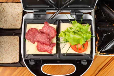 Téléchargez les photos : Une machine à sandwich avec deux sandwichs différents avec tomates, jambon, oignons et fromage. Préparation d'un repas léger. - en image libre de droit