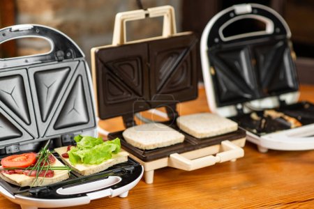 Photo for A variety of sandwiches that can be prepared in a sandwich maker concept. Row of electric sandwich makers on a wooden background. - Royalty Free Image