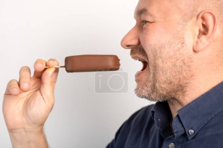 Foto de Hombre de mediana edad sin afeitar come helado en palo cubierto de chocolate sobre fondo blanco - Imagen libre de derechos
