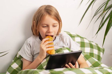 A young boy lounges on the bed, engrossed in his tablet, snacking on a pastry