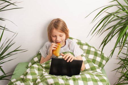 Seated on the bed, a boy unwinds with a tablet, enjoying a pastry. Bed rotting
