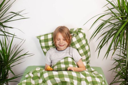 Bed rotting. Resting on the bed, a boy spends his day with a tablet, munching on a pastry
