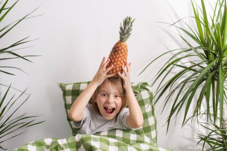 The young boy, about 9-10 years old, lies in bed, his long hair flowing, cradling a pineapple, symbolizing leisure and a carefree spirit