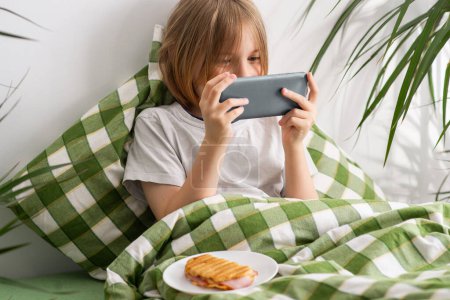 A young boy lies in bed looking at his phone and eating a sandwich