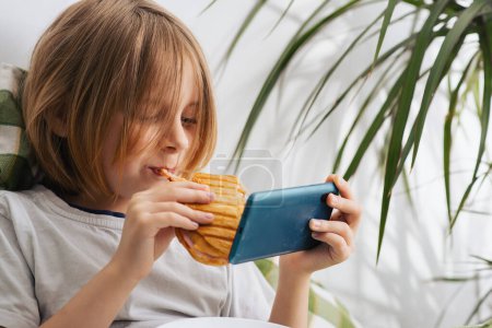 In the bed, a 9-10-year-old boy with lengthy hair savors a toast, leisurely using his mobile phone, exploring the digital realm, and gaming. Embodying the tranquility of relaxation and doing nothing