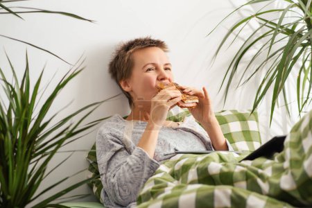 Engulfed in her tablet, a middle-aged woman with short hair spends the day in bed, eating a sandwich. Lethargy, lack of motivation. Bed rotting