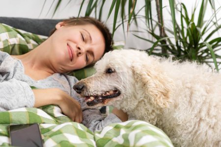 Throughout the day, a middle-aged woman with short hair relaxes in bed with her large poodle. Inactivity, weekend relaxation
